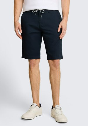 Mann trägt dunkelblaue Shorts mit weißem Kordelzug, weiße Sneakers mit schwarzen Details und ein weißes, hineingestecktes Hemd.