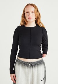 Frau mit roten Haaren, die ein schwarzes Langarmoberteil und graue Jogginghosen trägt, steht mit sichtbarem linken Arm und fehlendem rechten Arm unterhalb des Ellenbogens.