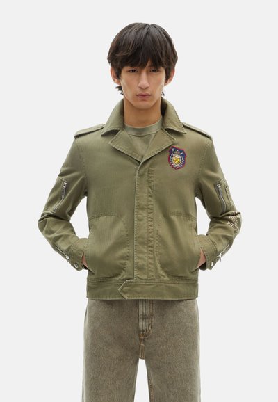 Olivengroene militaire stijl jas met een badge, voorkruiden, en met rits afgesloten mouwen, gemaakt van stevig materiaal met een gladde textuur.
