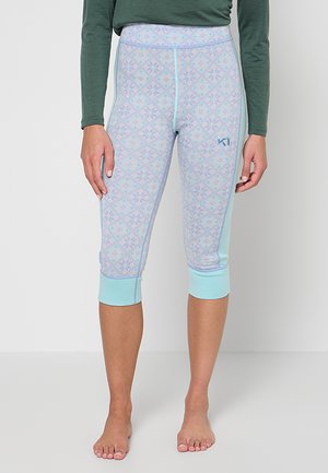 Frau trägt hellblaue Capri-Leggings mit Muster und ein dunkelgrünes langärmeliges Oberteil, steht barfuß vor einem schlichten Hintergrund.