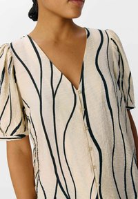 Blouse beige clair avec des lignes ondulées noires, décolleté en V, manches courtes bouffantes et boutons sur le devant. Tissu doux et texturé avec une coupe décontractée.