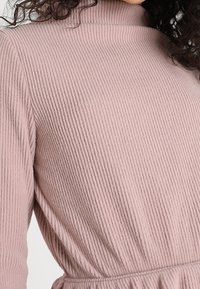 Gerippter, weicher rosa Pullover mit hohem Kragen und langen Ärmeln, der einen gerafften Bund für eine taillierte Silhouette aufweist.