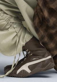 Botas altas marrones con un acabado texturizado, que presentan acentos en beige y una suela robusta. Combinadas con pantalones de un suave tono verde claro y texturizados.