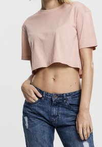 Persona che indossa una maglietta cropped rosa chiaro e jeans blu strappati, con una mano in tasca e l'altra appoggiata al fianco.