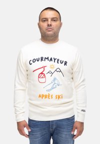 Maglione bianco lavorato a maglia con ricami colorati che raffigurano una scena di sci, montagne e il testo "COURMAYEUR APRES SKI". Jeans blu.