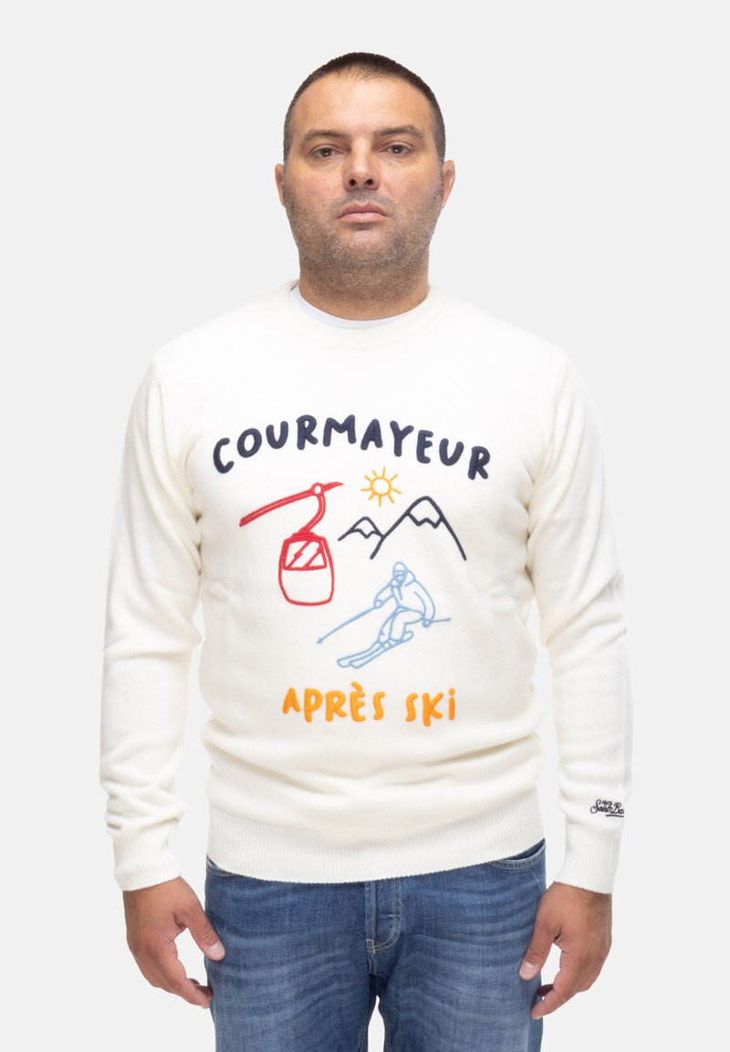 Maglione bianco lavorato a maglia con ricami colorati che raffigurano una scena di sci, montagne e il testo "COURMAYEUR APRES SKI". Jeans blu.