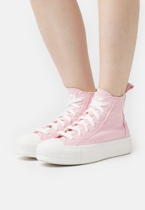 Fødder iført højt pink lærreds sneakers med hvide snørebånd og tykke hvide gummisåler på en helt hvid baggrund.