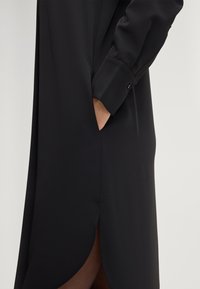 Zwarte jurk met lange mouwen en een soepele textuur, voorzien van een zijzak en een afgeronde zoom voor een slanke silhouet.