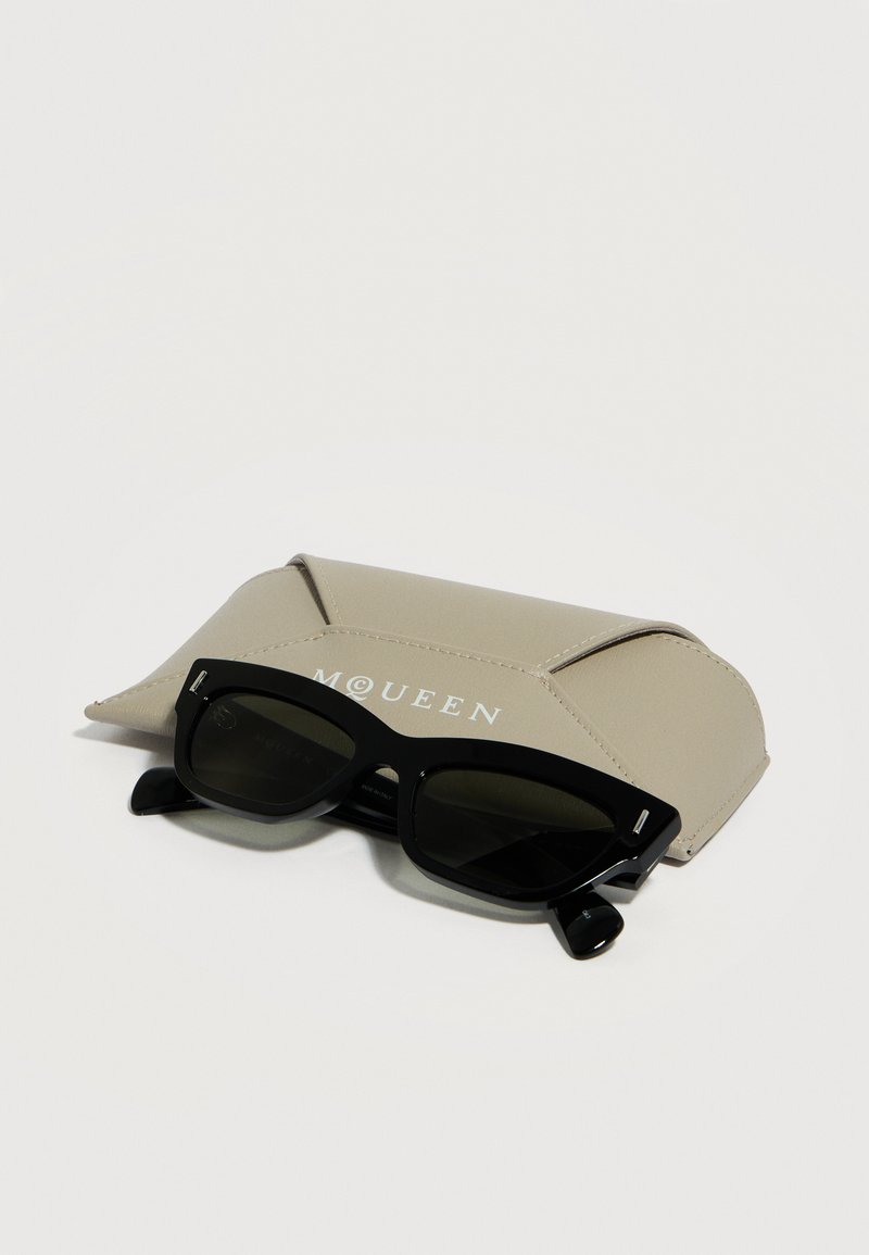 Schwarze rechteckige Sonnenbrille, die auf einem beigen Lederetui mit McQueen-Logo vor weißem Hintergrund liegt.