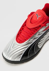 Scarpa sportiva rossa e argentata con un accento nero lucido, caratterizzata da una tomaia sintetica liscia, lacci testurizzati e un badge del logo in evidenza.