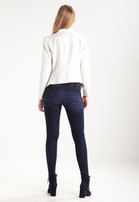 Femme debout de dos portant un blazer blanc, un jean skinny bleu marine, des bottines noires et une chemise bleu marine à pois blancs.