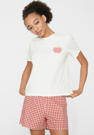 Femme aux cheveux bouclés portant un t-shirt blanc avec un motif de pomme rouge et un short à carreaux rouge et blanc, posant avec les mains sur les hanches.