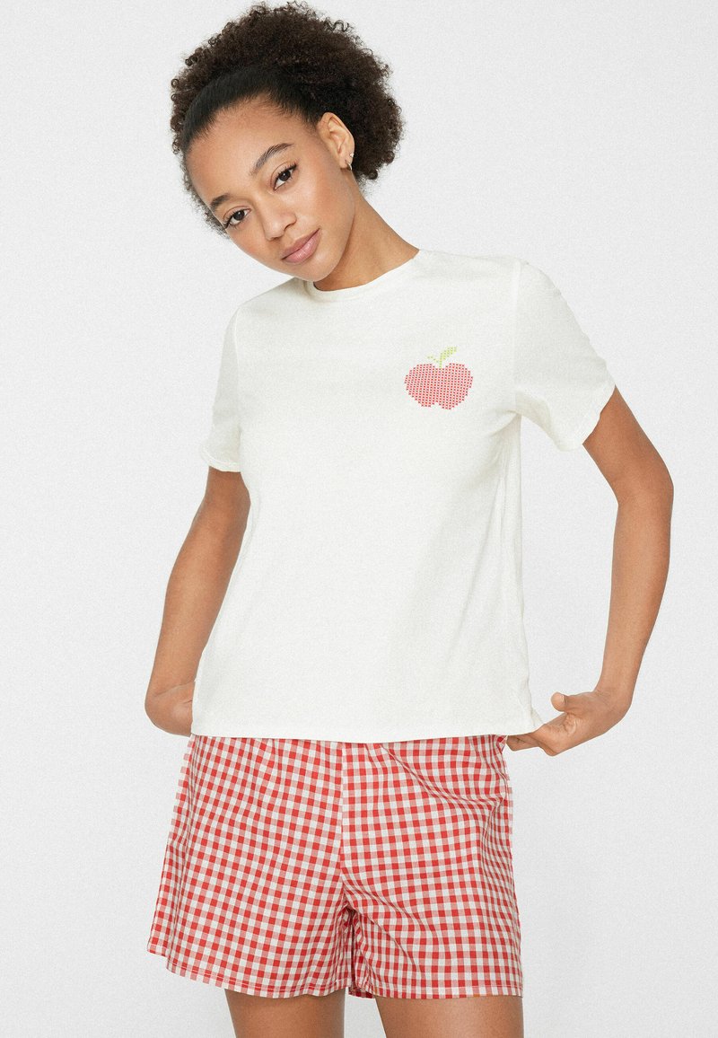 Frau mit lockigem Haar, die ein weißes T-Shirt mit einem roten Apfel-Design und rot-weiß karierte Shorts trägt, posiert mit den Händen in der Hüfte.