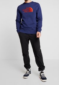 Man i blå The North Face-tröja, svarta joggerbyxor och navyvita sneakers, stående mot en enkel ljus bakgrund.