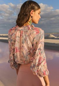 Floral, sheer blouse with ruffled collar and sleeves in light pink, featuring blue and metallic accents. Worn with fitted pink pants.