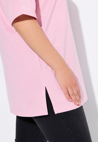 Hellrosa Oversized-T-Shirt mit kurzen Ärmeln und seitlichen Schlitzen, kombiniert mit schwarzen, eng anliegenden Jeans. Die weiche Stofftextur ist sichtbar.