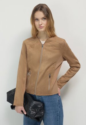 Junge Frau in einer sandfarbenen Jacke mit Reißverschluss und blauer Jeans, die eine große schwarze Leder-Clutch vor einem schlichten weißen Hintergrund hält.