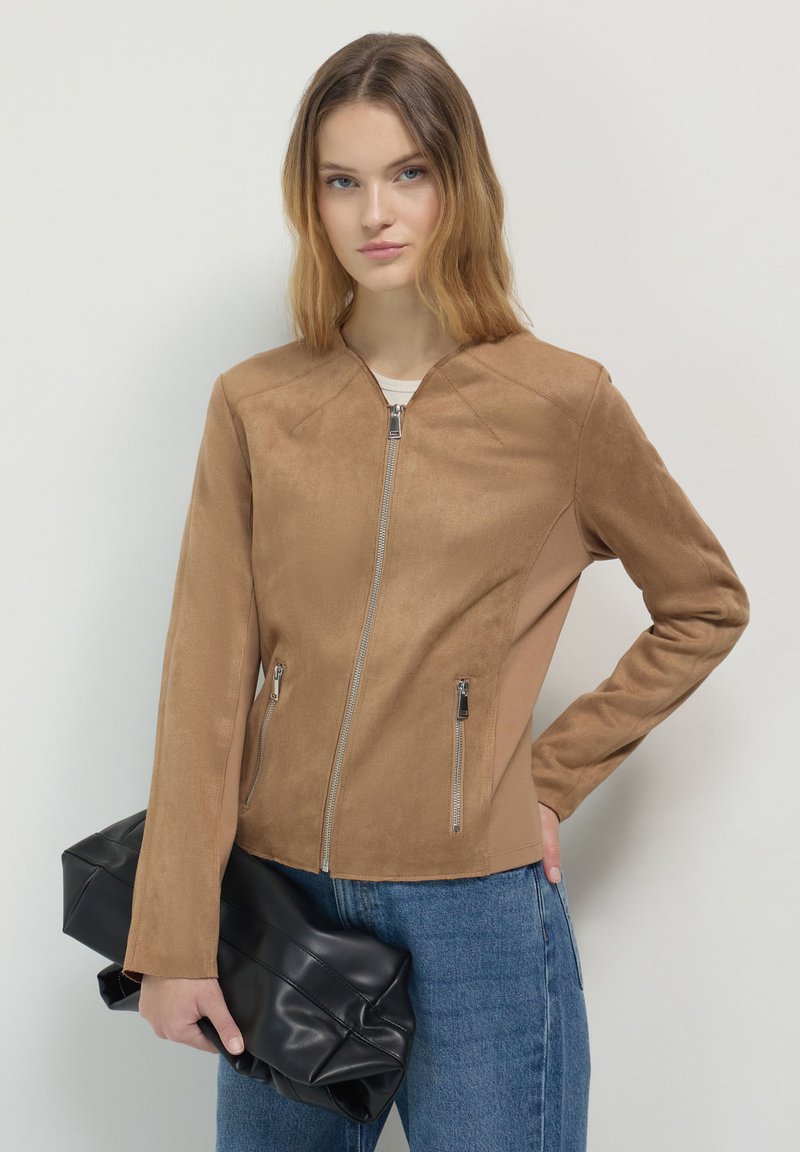 Giovane donna che indossa una giacca zip beige e jeans blu, mentre tiene una grande pochette in pelle nera contro uno sfondo bianco semplice.