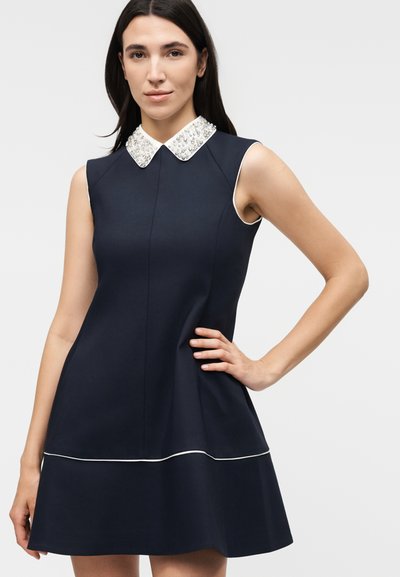 Γυναίκα με μακριά σκούρα μαλλιά που φοράει αμάνικο σκούρο μπλε φόρεμα με λευκή κεντημένη γιακά και λευκή διακοσμητική τρέσα κοντά στο τελείωμα, με το χέρι στη μέση.