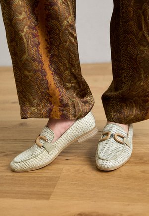 Tkané světle zelené loafers s kulatou špičkou, dřevěnou přezkou a nízkým dřevěným podpatkem, doplněné o vzorované hnědé kalhoty.