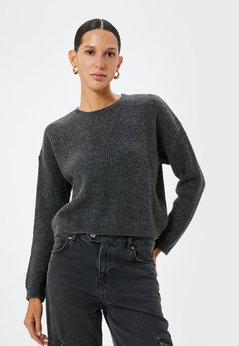 Grau gestrickter Pullover mit strukturiertem Rippmuster, Rundhalsausschnitt und langen Ärmeln, kombiniert mit dunklen Jeans. Verfügt über eine lässige Passform und dezente Details.