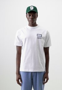 Hvit bomullst-skjorte med en liten svart og hvit logo på brystet, kombinert med lyseblå mønstrede shorts og en grønn caps.