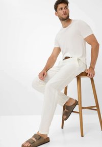 T-shirt bianco in cotone, pantaloni in lino color crema e sandali in suede marrone con cinturini larghi, seduto su uno sgabello di legno contro uno sfondo neutro.