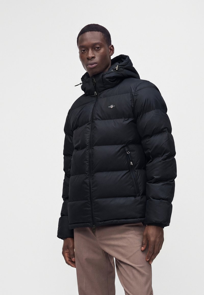 Homme portant une doudoune à capuche noire avec poches zippées et pantalon marron clair sur fond uni.