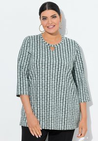 Donna sorridente con capelli scuri, indossa un top grigio e nero a fantasia con maniche a tre quarti e grandi orecchini a cerchio dorati.