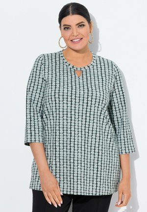 Donna sorridente con capelli scuri, indossa un top grigio e nero a fantasia con maniche a tre quarti e grandi orecchini a cerchio dorati.