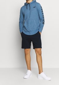 Blå långärmad huvtröja med svart logotyp och text på ärmen, kombinerad med mörka shorts och vita sneakers. Avslappnad atletisk stil.