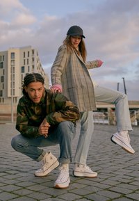 Białe sportowe buty za kostkę z kontrastującą gumową podeszwą, noszone przez osoby w casualowych stylizacjach, w tym w kurtkę w kratę i camo.