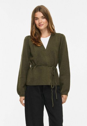 Femme portant un pull vert olive à manches longues et col en V avec une ceinture à la taille par-dessus un top blanc et un pantalon noir, debout devant un fond uni.