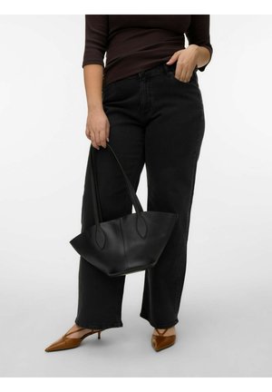 Person som bærer mørke jeans og brune hæler, holder en svart skinnveske i håndtakene mot en ensfarget hvit bakgrunn.