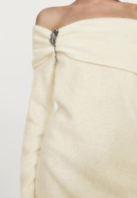 Cremefarvet, off-shoulder sweater lavet af blødt stof, med en sølvfarvet dekorativ broche formet som et blad på venstre skulder.