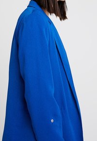 Jasnoniebieska marynarka wykonana z fakturowanej tkaniny, o dopasowanym kroju, z pojedynczym guzikiem przy mankiecie i wyciętymi klapami.