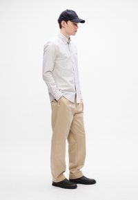 Gestreept overhemd met knopen in wit en grijs, beige broek, zwarte pet en zwarte schoenen. Heeft lange mouwen, een relaxte pasvorm en zijzakken.
