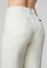 Pantalones de mezclilla blanca con un acabado texturizado, que presentan dos bolsillos traseros, una cintura alta y un diseño minimalista. Pequeña etiqueta de logo en la parte trasera.