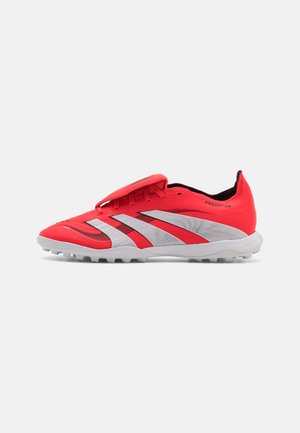 Chaussures de sport rouges avec un dessus texturé et une sangle réglable. Présentent trois rayures blanches et une semelle extérieure grise avec des crampons.