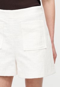 Λευκό σορτς από υφασμάτινο ύφασμα με ανάγλυφη υφή και μεγάλες εμπρόσθιες τσέπες, φορεμένο με σκούρο ριμπ τοπ, εμφανίζεται από τη μέση έως τη μέση του μηρού.