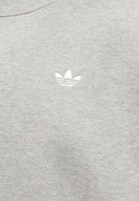 Tela gris clara con pequeño logo blanco bordado de Adidas Trefoil cerca del escote, mostrando textura y detalles de costura.