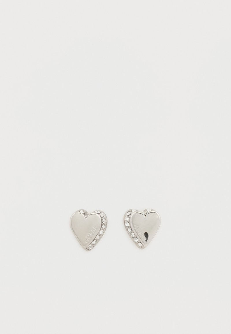 Boucles d'oreilles en forme de cœur argenté avec des accents en strass le long du bord. Finition polie avec le texte gravé "GUESS" sur la surface.