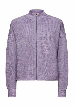 Personne portant une chemise longue bleu clair sous un pull court en tricot lavande à fermeture éclair, associé à un jean bleu foncé et un bracelet en or.