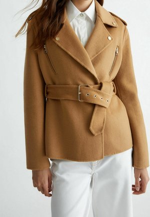 Femme portant une veste en laine couleur chameau ceinturée par-dessus une chemise blanche à col et un pantalon blanc, avec des détails zippés sur la poitrine.