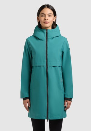 Kobieta w morskim, wodoodpornym płaszczu z kapturem, zapinanym na zamek, stojąca z ramionami opartymi swobodnie na tle jednolitego szarego tła.