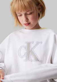 Biela mikina so širokým, štruktúrovaným strieborným logom "CK". Dlhé rukávy s rebrovanými manžetami, voľný strih a okrúhly výstrih.
