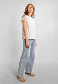 Top bianco con maniche corte in pizzo, abbinato a jeans a gamba larga blu chiaro e sandali beige. Texture liscia, vestibilità casual e design minimalista.