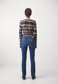 Persona con capelli corti che indossa orecchini a cerchio, una maglietta a maniche lunghe nera e grigia tie-dye, jeans blu e scarpe nere, girata di spalle.