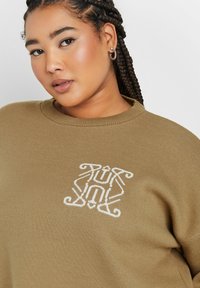 Olivgrüner Strickpullover mit einem weißen gestickten Design auf der Brust und einem Rundhalsausschnitt. Weiche Textur mit gerippten Bündchen.