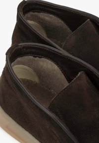 Brązowe zamszowe buty na slip-on z miękką podszewką, zaokrąglonym kształtem i kontrastowym stebnikiem. Charakteryzują się minimalistycznym wzornictwem i gładką podeszwą.